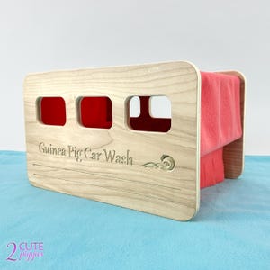 May include: A wooden Guinea Pig Car Wash with three red openings and a pink fleece curtain. The car wash is made of natural wood and has a carved design on the front.