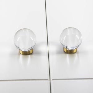 May include: Two clear, spherical cabinet knobs with a brass base are mounted on a white cabinet door. The knobs are transparent, allowing a view of the cabinet's surface. The brass base adds a touch of elegance to the hardware.