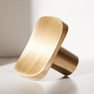 May include: A brushed gold square cabinet knob with a rounded edge and a short, cylindrical base.