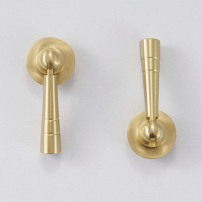 K&ouml;nnte beinhalten: Zwei goldfarbene Schrankgriffe mit runder Basis und einem langen, schlanken Griff. Die Griffe haben ein geriffeltes Design.
