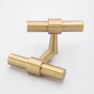 May include: Two brushed gold T-shaped cabinet knobs. The knobs have a cylindrical shape with a slightly rounded end. The knobs are arranged in a staggered position.