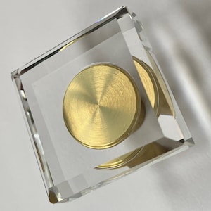 May include: A square, clear glass display case with a gold-colored round disc inside.
