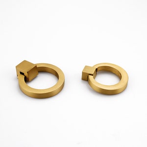 K&ouml;nnte beinhalten: Zwei goldfarbene Metall-Ringgriffe mit quadratischen Griffen. Die Ringgriffe sind eine beliebte Wahl f&uuml;r Schrankbeschl&auml;ge.
