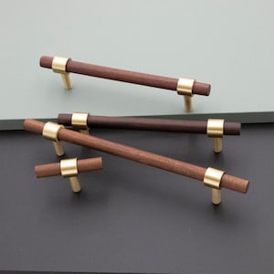 May include: Four wooden cabinet knobs with gold metal accents. The knobs are in various sizes and shapes, including a T-shaped knob and a long, rectangular knob.