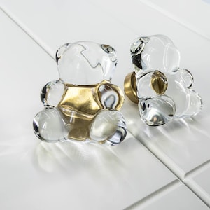 May include: Two clear glass bear-shaped cabinet knobs with gold accents. The knobs are designed to be used on furniture.