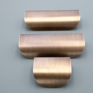 May include: Three brushed brass cabinet pulls in different sizes. The pulls are curved and have a rounded edge.