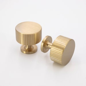 May include: Two gold-toned cabinet knobs with a ribbed design. The knobs are round and have a small base.