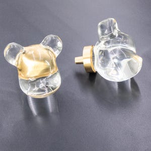 May include: Two clear glass cabinet knobs in the shape of a bear. One knob has a gold accent on the bottom half. Both knobs have a brass base.