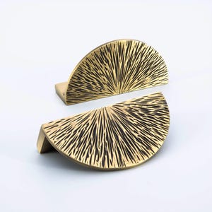 May include: Two brass-colored, semi-circular cabinet pulls. Each pull has a textured, sunburst-like pattern. The pulls are mounted on small, rectangular bases, and are ideal for adding a touch of elegance to furniture.