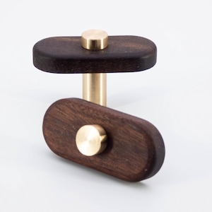 Peut inclure: Deux boutons de meuble en bois avec un corps ovale brun foncé et un bouton circulaire de couleur laiton. Les boutons sont reliés par une tige de couleur laiton. Les boutons sont posés sur un fond blanc.