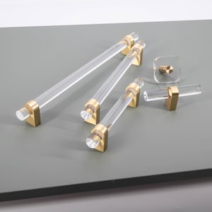 May include: A selection of clear acrylic cabinet handles and knobs with gold-coloured metal detailing. The handles are cylindrical, and the knobs are square and T-shaped. The hardware is displayed on a grey surface.