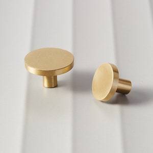 May include: Two brass cabinet knobs with a circular design. The knobs have a flat, disc-shaped top and a cylindrical stem. The finish is a warm, golden color, and the knobs are set against a neutral background.