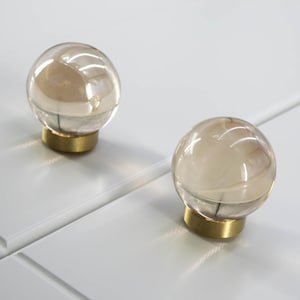 May include: Two clear glass ball cabinet knobs with a gold-colored base. The knobs are round and transparent, reflecting light. They are mounted on a white surface, likely a cabinet door or drawer front. The knobs add a touch of elegance.