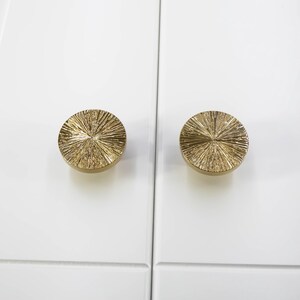 Puede incluir: Dos tiradores dorados para armario con un dise&ntilde;o texturizado de rayos de sol. Los tiradores redondos est&aacute;n montados en una puerta de armario blanca. Los tiradores son de metal y tienen un acabado brillante.