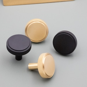 May include: Four cabinet knobs in gold and black. Two gold knobs have a textured, knurled edge and a flat top. The other two knobs are black, one with a textured edge and a flat top, and the other with a smooth top.