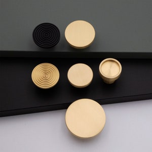 May include: Five cabinet knobs, four are gold and one is black. The gold knobs are round and have a smooth finish. The black knob is round and has a concentric circle design.