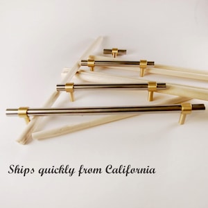 May include: A collection of cabinet pulls with a modern design. The pulls feature a brushed metal bar with gold-toned accents. The pulls are arranged on a white surface with wooden dowels.