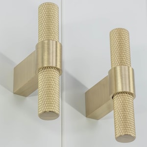 May include: Two brass-colored cabinet pulls. Each pull features a cylindrical handle with a diamond pattern, connected to a rectangular base. The pulls are mounted on a white surface, showcasing a modern design.