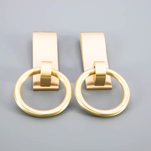 May include: Two brass-colored cabinet pulls with a rectangular backplate and a circular ring handle. The pulls are made of metal and have a smooth, matte finish. The background is a solid gray color.