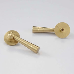 K&ouml;nnte beinhalten: Zwei goldfarbene Schrankkn&ouml;pfe mit runder Basis und einem langen, zylindrischen Griff. Die Griffe haben ein gerilltes Design.