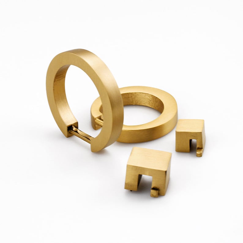 K&ouml;nnte beinhalten: Ein Satz aus drei goldfarbenen Metall-Schrankgriffen. Zwei der Griffe sind runde Ringe, und einer ist ein Quadrat mit einem Ausschnitt in der Mitte.