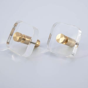 May include: Two clear acrylic wall hooks with gold metal hooks. The hooks are square shaped and have a modern design.