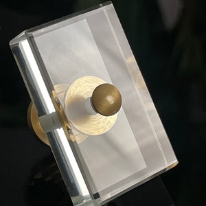 May include: A clear glass rectangular box with a brass knob on top. The box is frosted and has a subtle sheen.