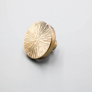 Puede incluir: Un pomo de armario redondo dorado con un dise&ntilde;o texturizado de rayos de sol. El pomo tiene un peque&ntilde;o v&aacute;stago cil&iacute;ndrico para la fijaci&oacute;n. Este herraje decorativo es adecuado para cajones, armarios y muebles.