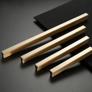 May include: Four gold-toned metal cabinet pulls with a rectangular shape and squared edges. The pulls are arranged in a staggered pattern on a black surface.