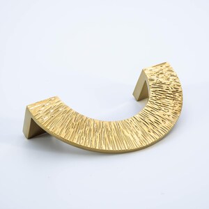 Puede incluir: Un tirador de armario dorado, semicircular, con un patr&oacute;n lineal texturizado. El tirador tiene un asa curva y dos postes de montaje rectangulares. El herraje est&aacute; dise&ntilde;ado para muebles y decoraci&oacute;n del hogar.