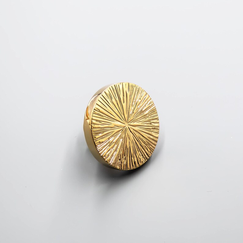 Puede incluir: Un pomo de armario circular dorado con un dise&ntilde;o de rayos de sol. El pomo tiene una superficie texturizada con l&iacute;neas radiales, creando un acento decorativo y elegante para muebles. El herraje es de metal.