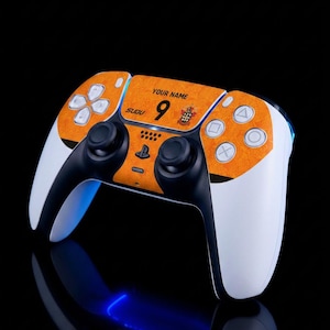 Può includere: Un controller di gioco wireless bianco e nero con una pellicola testurizzata arancione sulla parte frontale. La pellicola mostra il testo "YOUR NAME", il numero "9", la parola "SUDU" e un piccolo stemma. Il controller ha pulsanti standard.