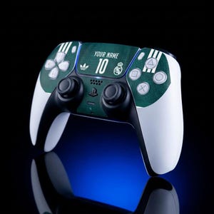 Puede incluir: Un mando de juego blanco y negro con una cubierta texturizada de color verde oscuro en el panel central. El diseño incluye el texto YOUR NAME, el número 10, un logotipo de Adidas y el escudo del Real Madrid. Tiene botones y joysticks estándar.