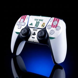 May include: A white and black wireless video game controller featuring a custom graphic design with the text YOUR NAME, the number 10, a Real Madrid club crest, and three red stripes on the side panels, alongside standard black analog sticks and buttons.