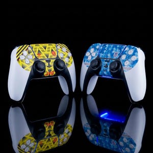 May include: Two wireless game controllers with custom skins. The left controller features a yellow and black geometric pattern with an Adidas logo and Arsenal crest. The right controller has a blue and white mosaic pattern with an England football crest.