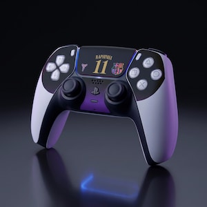May include: A black and white wireless game controller featuring a purple center panel, the text RAPHINHA 11, a small logo of a basketball player, and the FC Barcelona crest printed on the central touchpad area with standard gaming buttons.