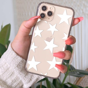 May include: Transparent phone case featuring a white star pattern. The phone is held in a hand with long, pink nails. The phone has a triple-lens camera.