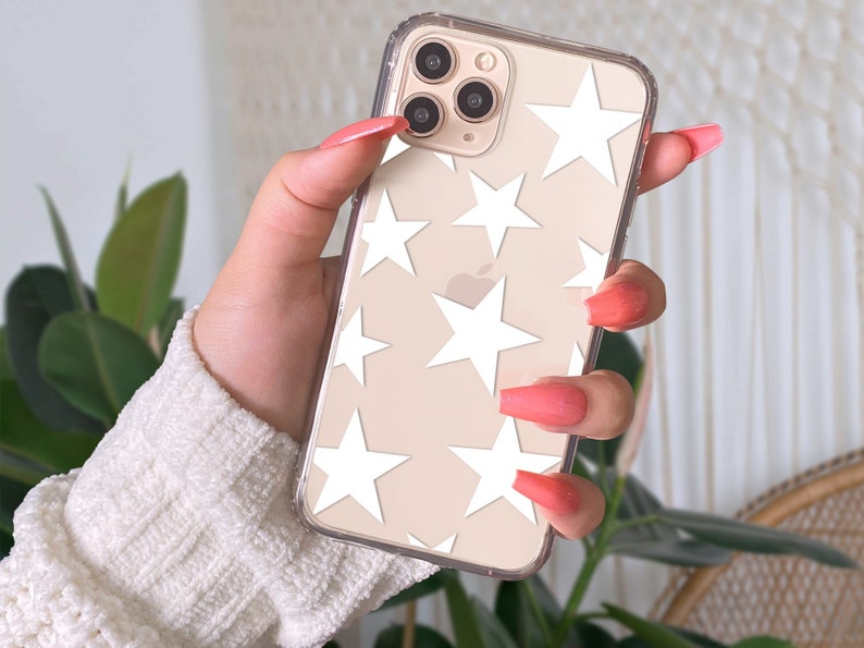 May include: Transparent phone case featuring a white star pattern. The phone is held in a hand with long, pink nails. The phone has a triple-lens camera.