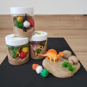 May include: Three clear jars filled with brown sand and small toys, including dinosaurs, plants, and pom-poms. A separate pile of brown sand with a small orange dinosaur and green plants.