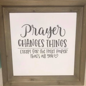 Op de afbeelding: Een ingelijst bord met de woorden "Prayer Changes Things" in een decoratief lettertype. Daaronder staat "Except for the toilet paper. That's all you ❤️". Het frame is lichtbruin.