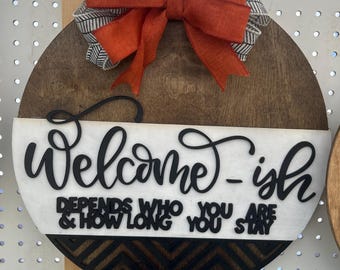May include: A round wooden sign with the text "Welcome-ish" and "Depends who you are & how long you stay." The sign has a decorative orange bow and a black and white chevron pattern.