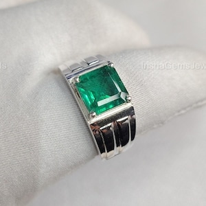 May include: A silver ring featuring a rectangular, emerald-cut green gemstone. The ring has a modern design with a polished finish and a band with vertical grooves. The ring is displayed on a white surface.