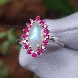 May include: A silver ring featuring a central, oval-shaped moonstone, surrounded by a halo of small, round, bright pink gemstones. The moonstone has a pearlescent, iridescent quality. The ring is set against a white background.