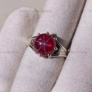 May include: A silver ring featuring a large, round, star ruby gemstone. The ruby is a deep red color with a six-rayed star pattern. The ring is held by a gloved hand.