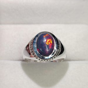 Könnte beinhalten: Ein silberner Ring mit einem ovalen, dunklen Edelstein mit leuchtenden roten und orangen Farbtönen. Der Ring hat ein detailliertes Band und wird in einer hellen Schachtel präsentiert. Der Text "InshaGemsJewels" ist sichtbar.