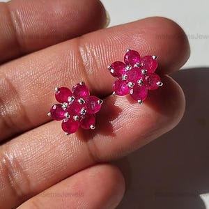 Pode incluir: Um par de brincos de botão em forma de flor. Cada brinco apresenta um aglomerado de pequenas pedras preciosas rosa brilhantes e redondas, cravejadas em metal prateado. Os brincos são exibidos em um fundo claro.