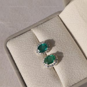 Può includere: Un paio di orecchini a lobo con smeraldi taglio smeraldo e un alone di piccole gemme trasparenti. Gli orecchini sono incastonati in un metallo color argento e sono esposti in un portagioie grigio.