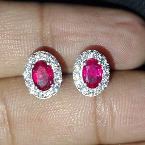 Puede incluir: Un par de pendientes de plata con piedras preciosas de rubí rojo de forma ovalada rodeadas por un halo de piedras blancas más pequeñas.