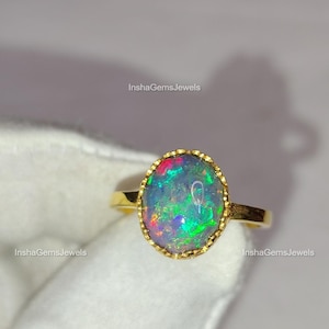May include: A gold ring featuring an oval opal gemstone with vibrant colors of green, blue, and red. The opal is set in a gold bezel with a textured edge. The ring is displayed on a white surface, with the text "InshaGemsJewels" visible.