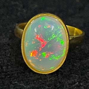 May include: A gold ring with an oval-shaped opal gemstone. The opal has a white base with flashes of red and green.